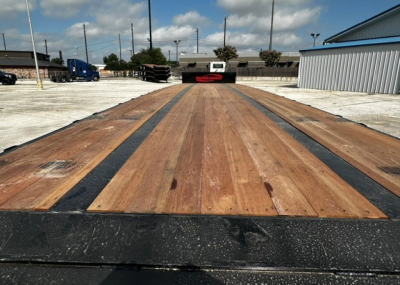 Flatbed trailer bed with wooden deck planks and black steel edge; distant trucks and industrial yard in background.