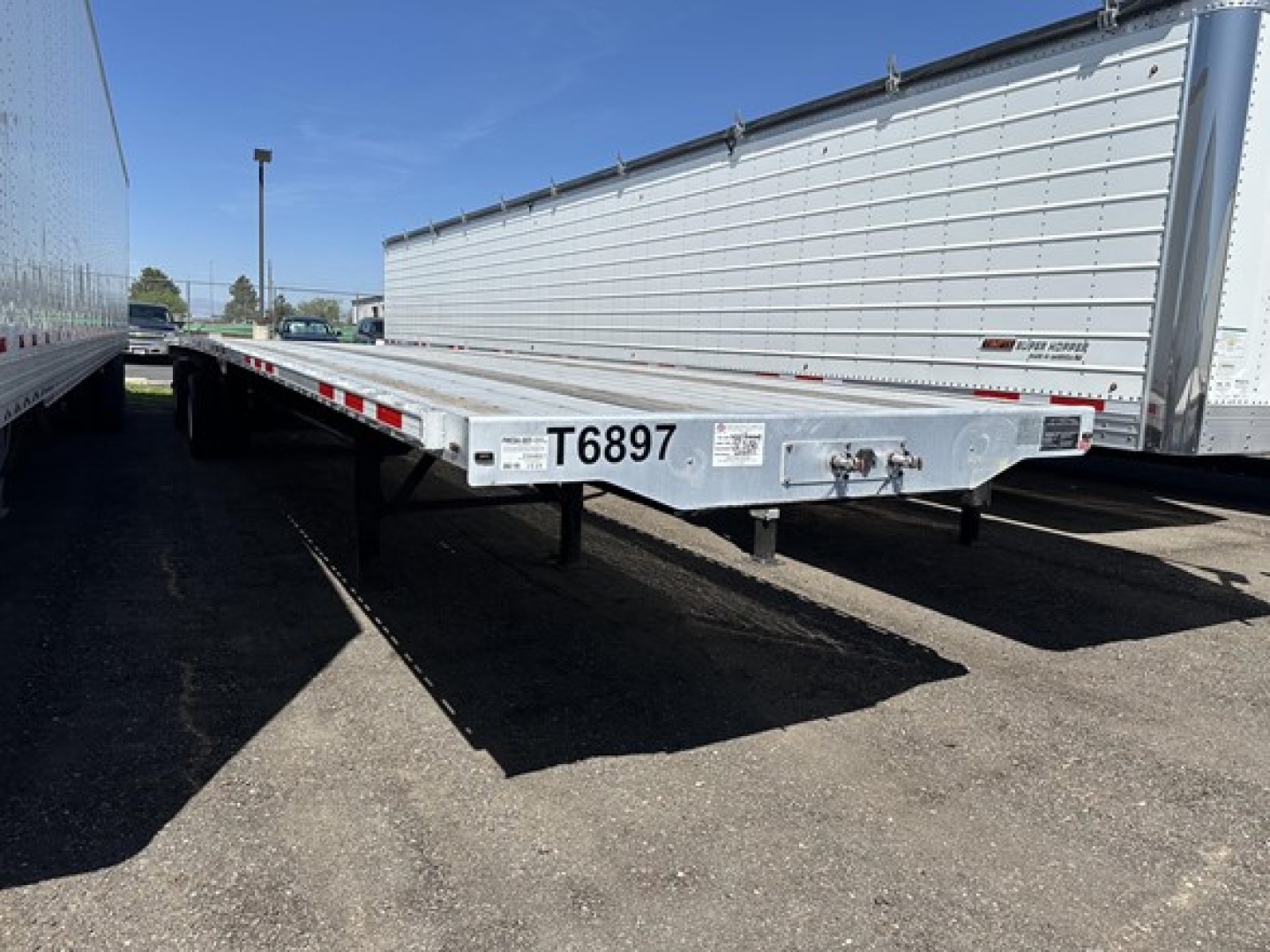 Flatbed trailer showing front end with T6897 printed on the tongue, low-angle view, other trailers in background