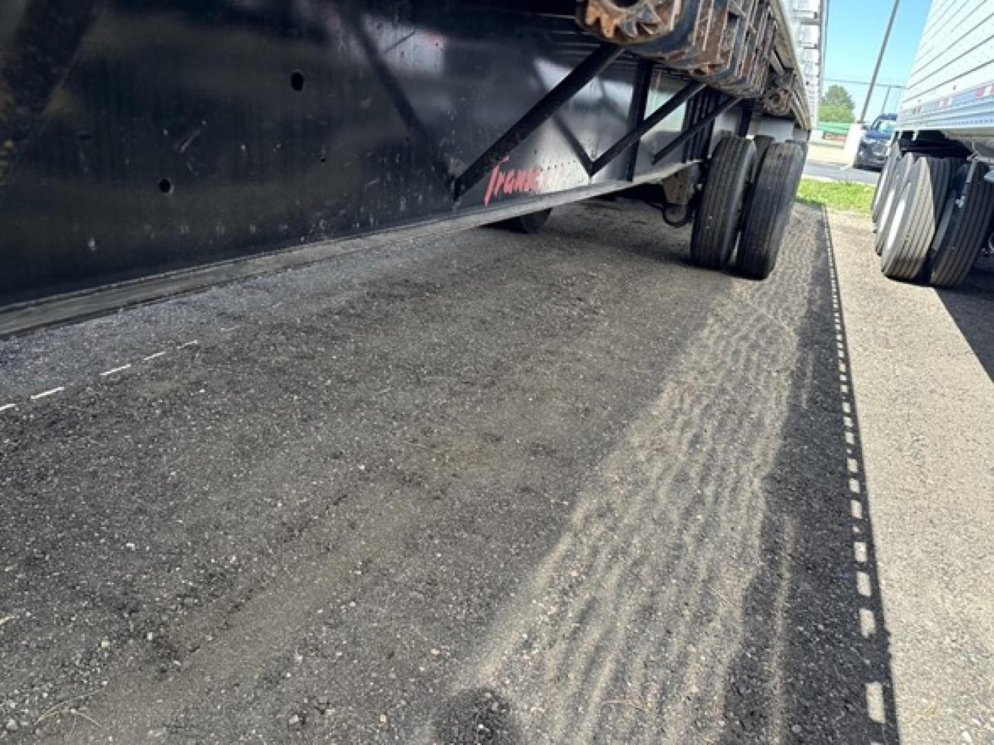 Flatbed trailer undercarriage showing wheel axles and deck edge, with reflective tape along the side; visible text reads Transcraft on the frame.