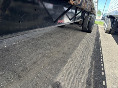 Flatbed trailer undercarriage showing wheel axles and deck edge, with reflective tape along the side; visible text reads Transcraft on the frame.