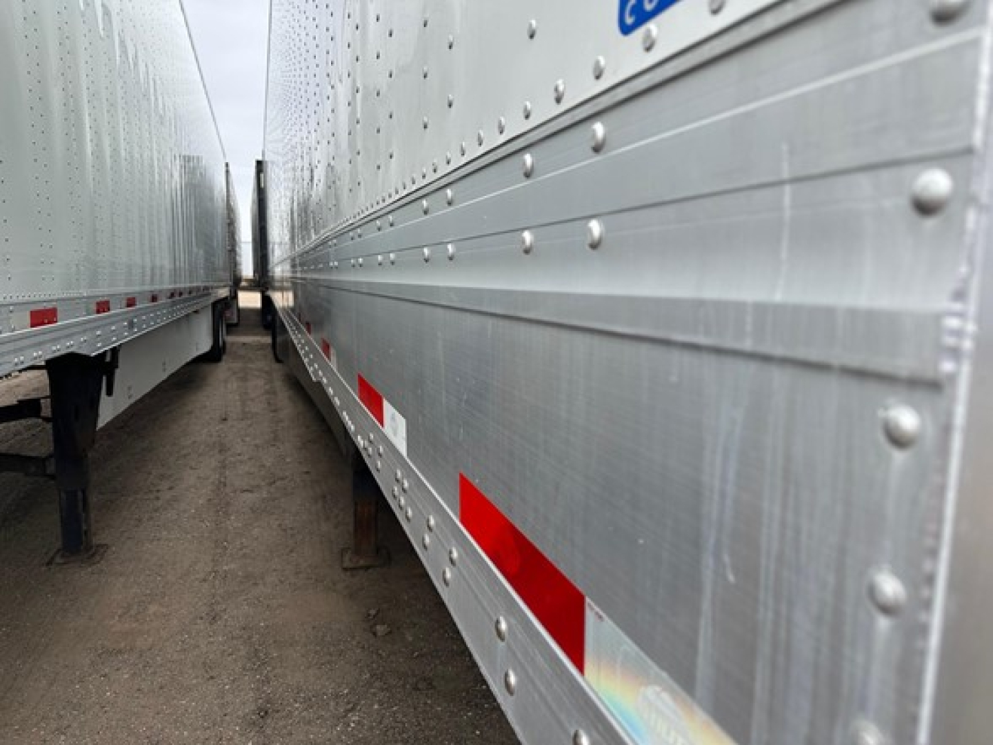 Trailer side view showing riveted aluminum paneling with reflective red/white side marker tape, underside supports, and a narrow passage between two trailers.
