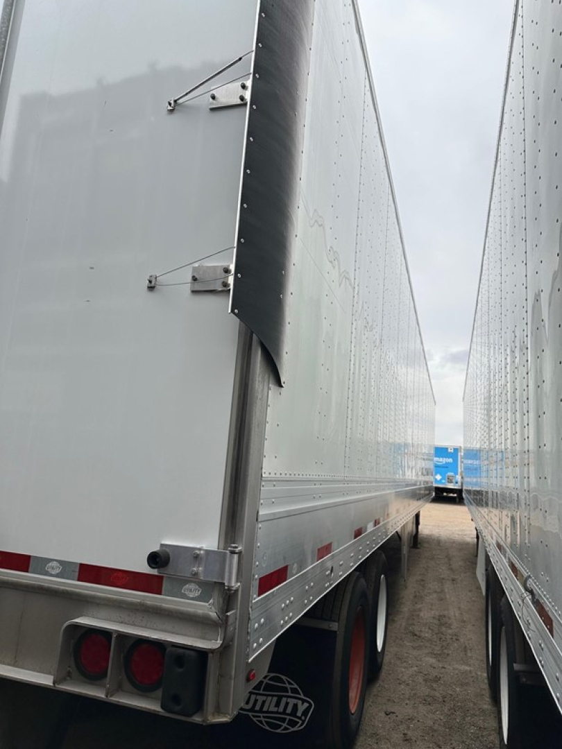 Trailer side view showing a Utility 4000Dx box trailer with riveted aluminum walls, loading seam, and rear light cluster; perspective from between two trailers.