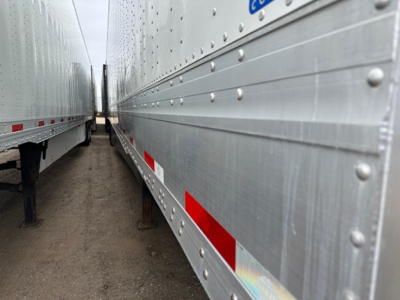 Trailer side view showing riveted aluminum paneling with reflective red/white side marker tape, underside supports, and a narrow passage between two trailers.