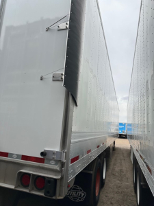 Trailer side view showing a Utility 4000Dx box trailer with riveted aluminum walls, loading seam, and rear light cluster; perspective from between two trailers.