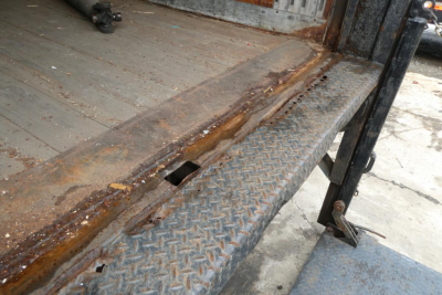 Truck bed edge with rust and diamond plate step; side view shows damage to metal platform and part of hydraulic lift.