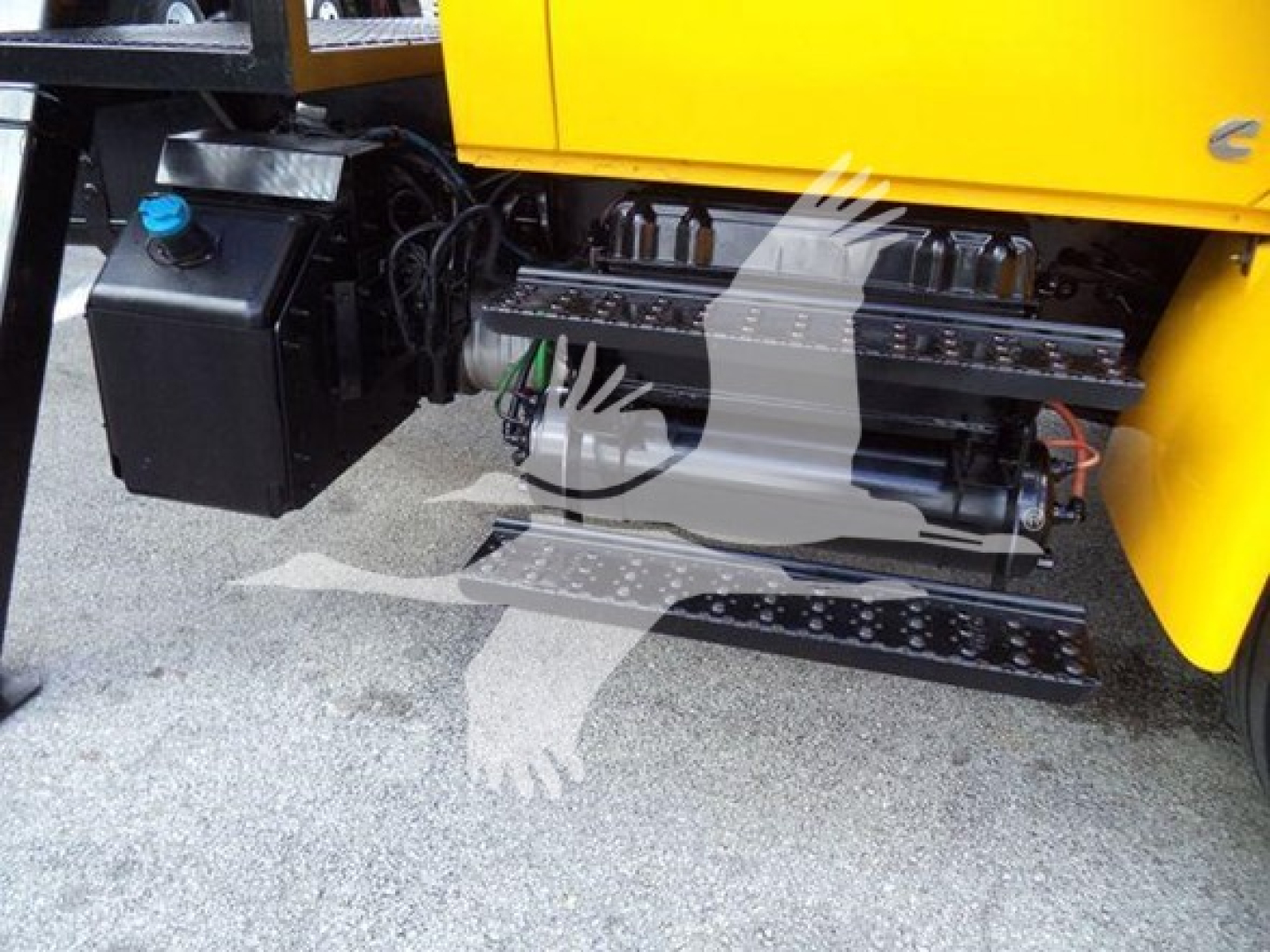 Truck side view showing fuel/fluids tank on left and mechanical components under a yellow dump body; visible hoses, lines, metal steps, and a black engine area.