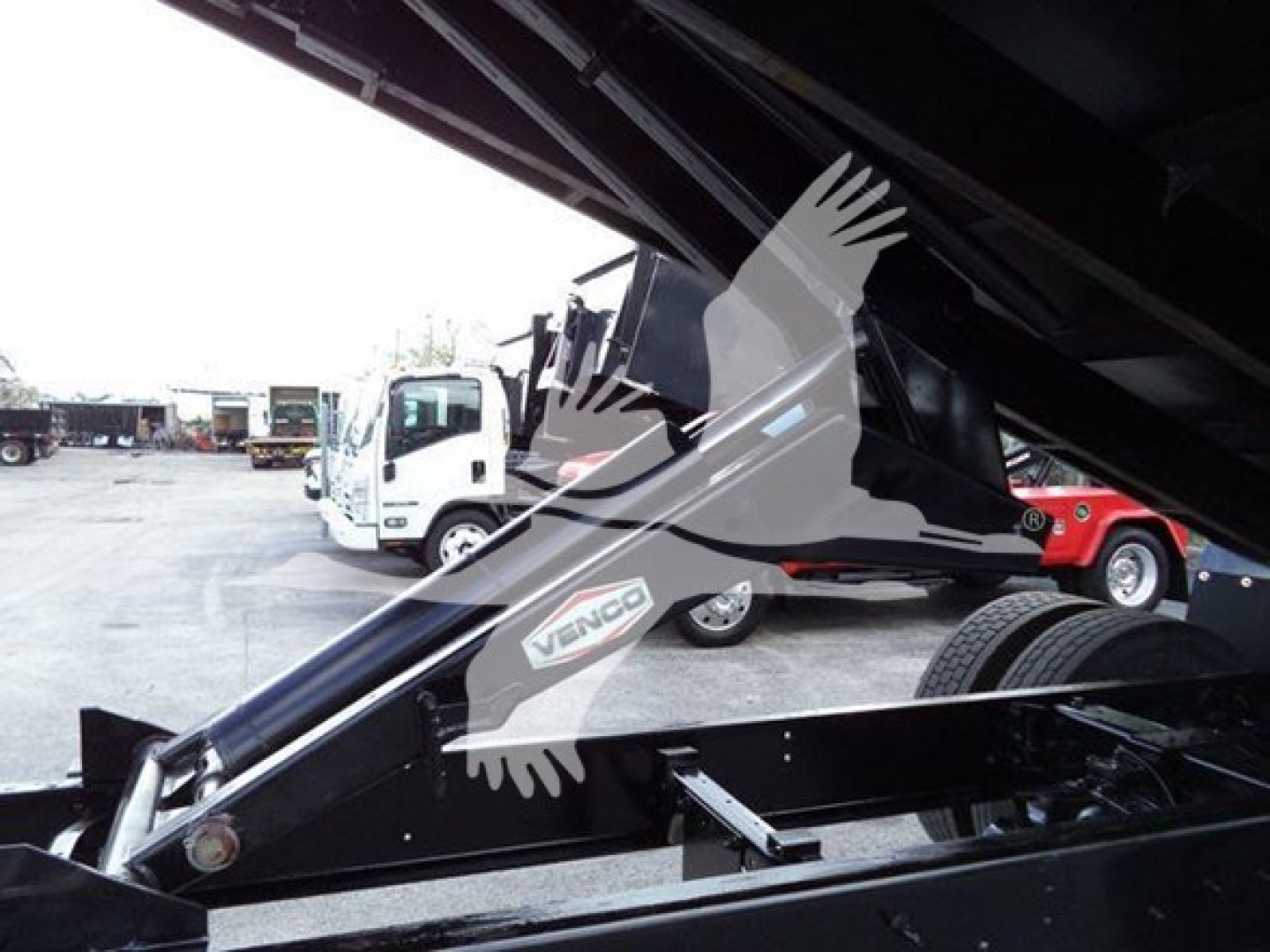 Grapple truck undercarriage with black metal framework and hydraulic arm; sticker shows 'VENGO' or 'VNGO' on side; background shows other trucks in a lot.