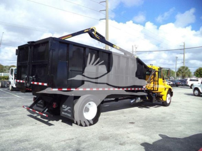 Truck bed with grapple-equipped dumpster body attached to a yellow loader, side view.