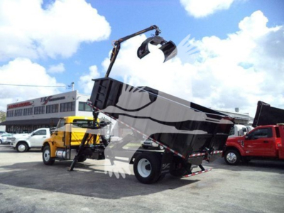 2019 International Durastar grapple truck with front-end loader bucket raised, black hydraulic arm, dump bed tilted, in yard among other trucks.