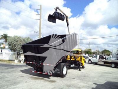 Dump trailer with grapple loading crane in robotic arm position over top. Large dump container shows silhouette graphic on side, rear wheels visible, clear sky.