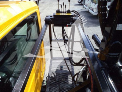 Grapple truck cab and hydraulic arms in the process of loading, with yellow vehicle on left and black equipment on right.