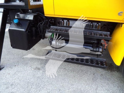 Truck side view showing fuel/fluids tank on left and mechanical components under a yellow dump body; visible hoses, lines, metal steps, and a black engine area.