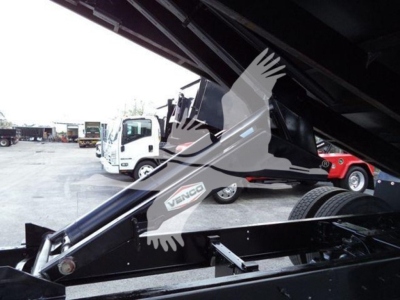 Grapple truck undercarriage with black metal framework and hydraulic arm; sticker shows 'VENGO' or 'VNGO' on side; background shows other trucks in a lot.