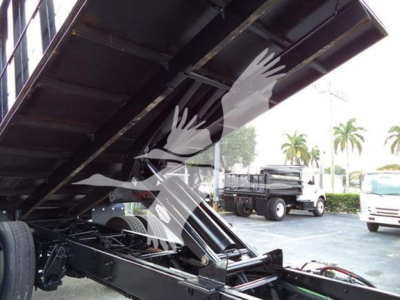 Truck bed in raised position showing grapple truck component and hydraulic lines with other trucks in background.