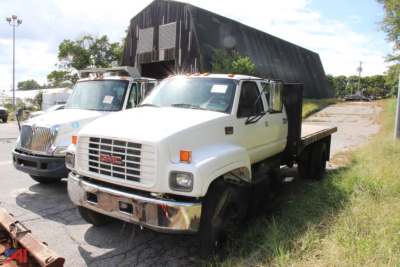 GMC medium-duty truck in a yard with a dump bed raised, two white flatbed trucks, GMC grille visible, VIN plate not legible.