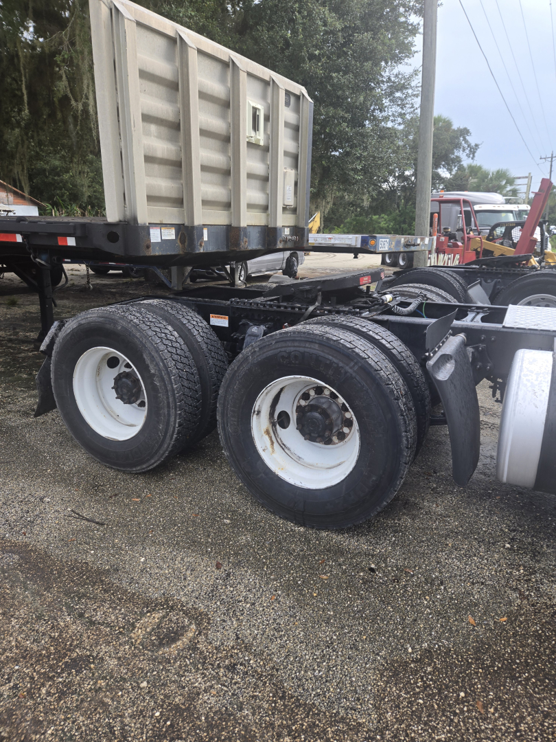 Truck chassis with tandem rear axles and dual wheels, Freightliner Cascadia Day Cab likely, with a large aluminum dump trailer in background.