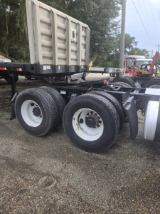 Truck chassis with tandem rear axles and dual wheels, Freightliner Cascadia Day Cab likely, with a large aluminum dump trailer in background.