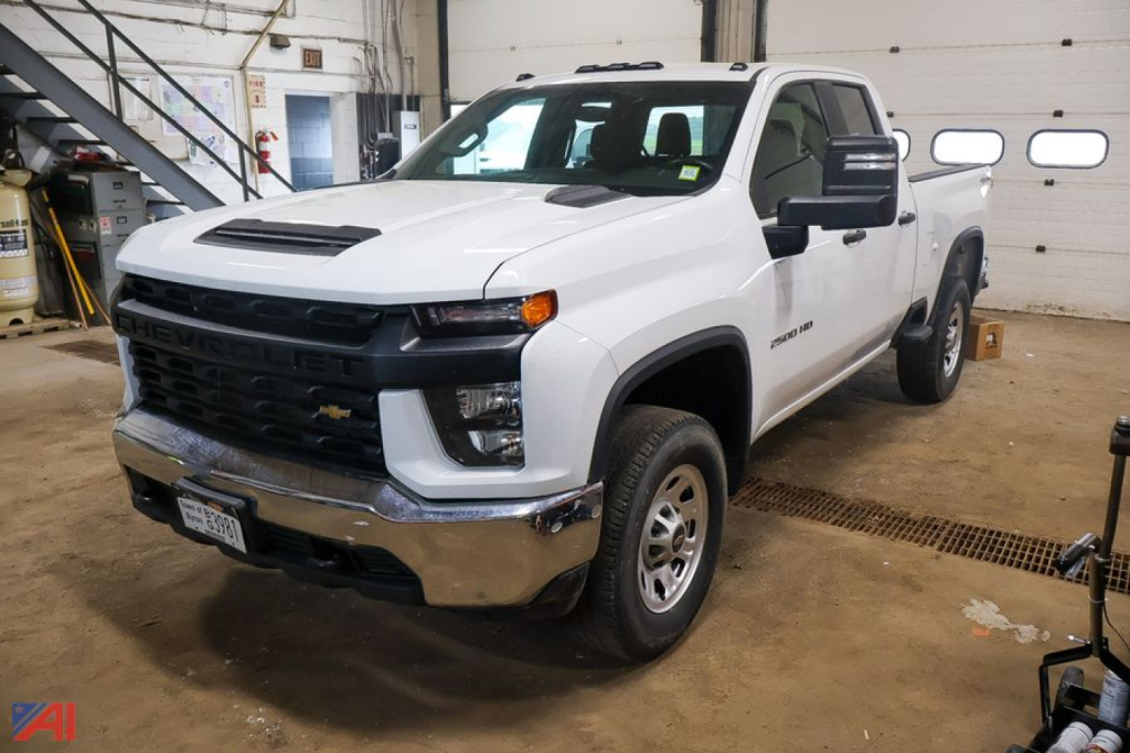 Front-left view of a white 2023 Chevrolet Silverado 2500HD Crew Cab pickup truck inside a shop. Visible text includes Chevrolet badge on grille and ‘ Silverado 2500HD ’ on side, license plate partial.