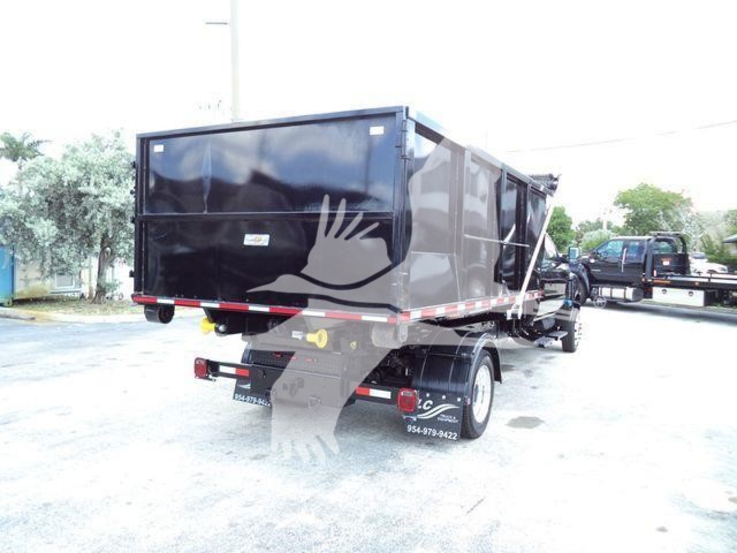 Dump truck/roll-off body rear view showing black container on a chassis with rear lights and dual wheels, side letters and contact numbers faintly visible.