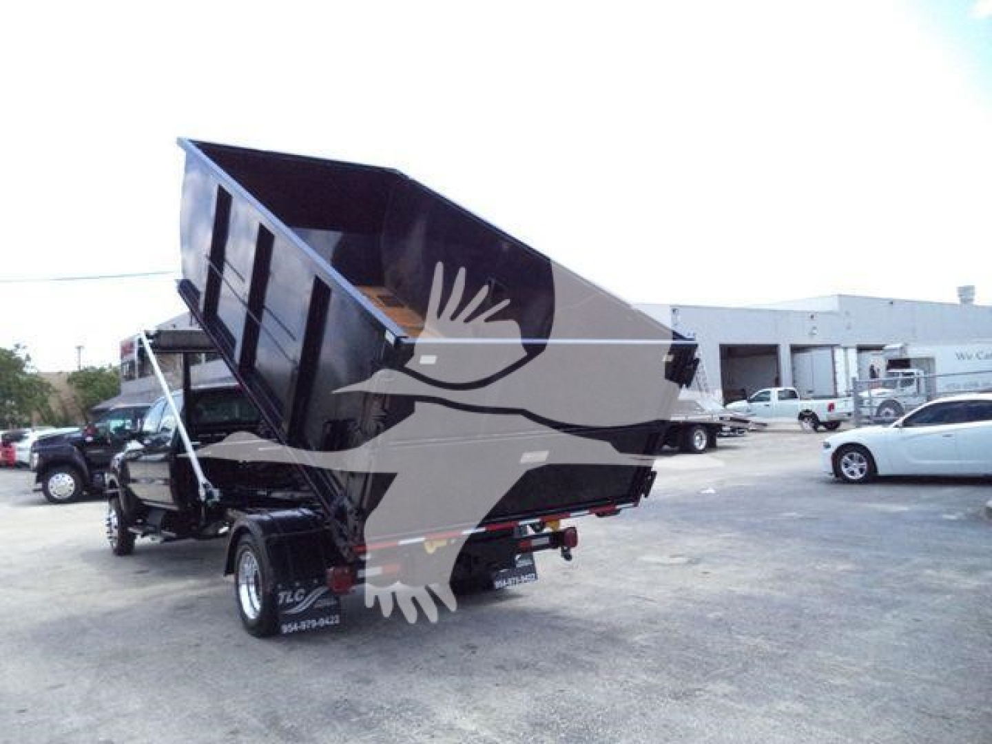 Dump truck tail with raised dump bed in a parking lot, with shadowed logo overlaid showing a bird in flight.