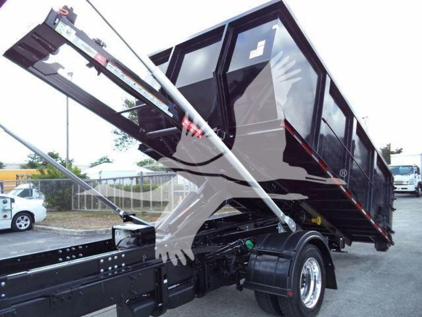 Image shows a black International CV roll-off truck body with a hydraulic arm and bed raised. Visible text/labels not clearly legible; includes side window and chassis components.