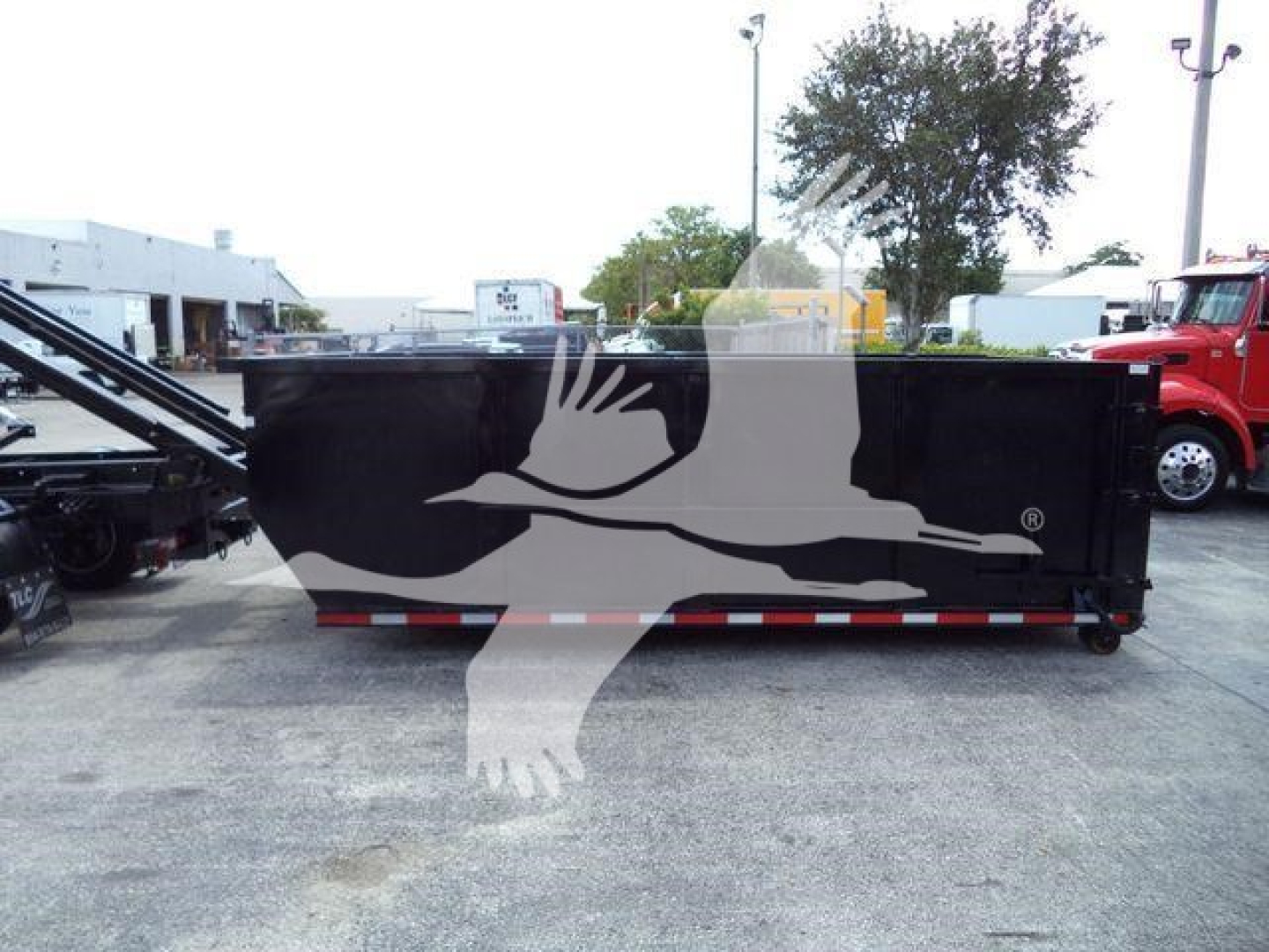 Rolloff truck body in a lot showing a large black dumpster with a faint white flying bird graphic overlay; red and white reflective strip along bottom edge; surroundings include other trucks and industrial buildings.