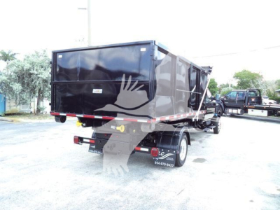 Dump truck/roll-off body rear view showing black container on a chassis with rear lights and dual wheels, side letters and contact numbers faintly visible.