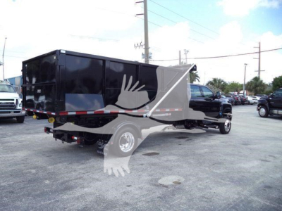 Truck in lot showing a black roll-off container on a heavy-duty chassis with rear hook lift and dual rear wheels.
