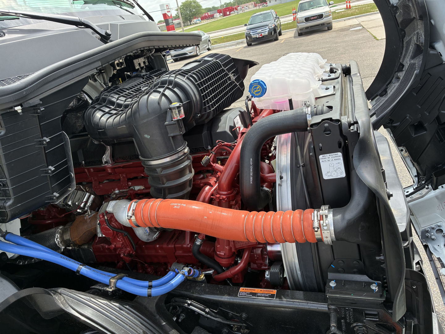 Engine bay of a 2027 Mack PR64T76H showing the red engine, black air intake, orange coolant hose, blue hoses, radiator, and coolant reservoir cap.