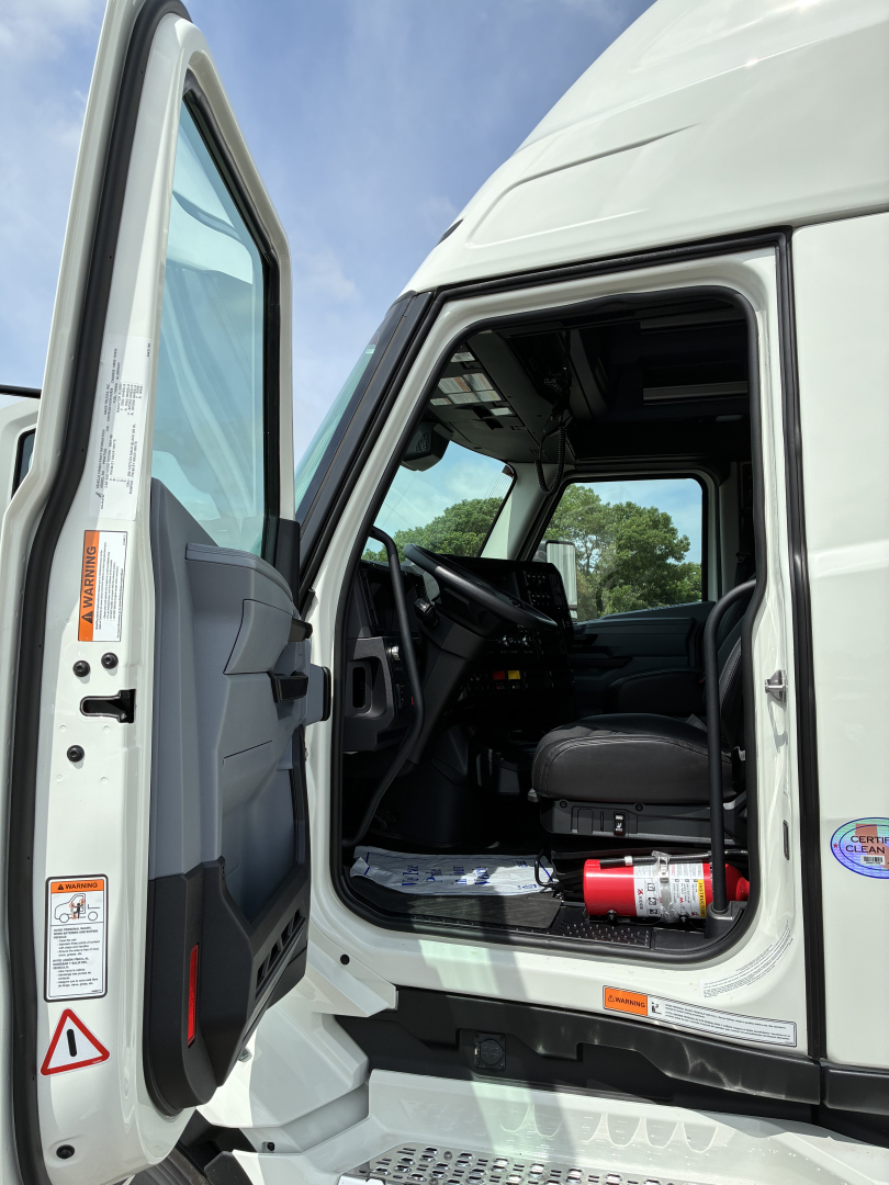 Truck cab interior with open door showing steering wheel, dashboard, and driver's seat; visible labels on door edge and step; small fire extinguisher mounted under dash.