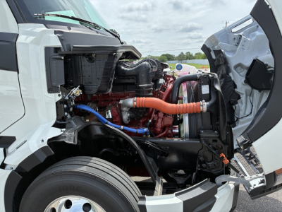 Truck engine bay of a 2027 Mack PR64T76H with orange coolant hose, blue braided line, red engine components and open hood side view.