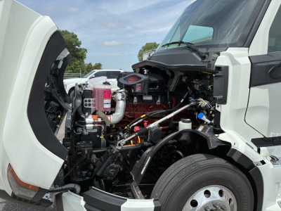 Truck engine bay with open hood showing red engine block, intake piping, hoses, and various components in a white Mack PR64T76H cab. 