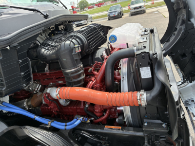 Engine bay of a 2027 Mack PR64T76H showing the red engine, black air intake, orange coolant hose, blue hoses, radiator, and coolant reservoir cap.