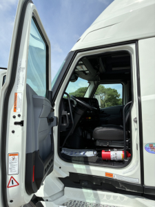 Truck cab interior with open door showing steering wheel, dashboard, and driver's seat; visible labels on door edge and step; small fire extinguisher mounted under dash.