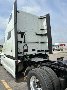 Truck cab rear with open door reveals interior wiring and chassis of a white Mack PR64T76H sleeper tractor. Side rear wheels and empty hitch area visible.