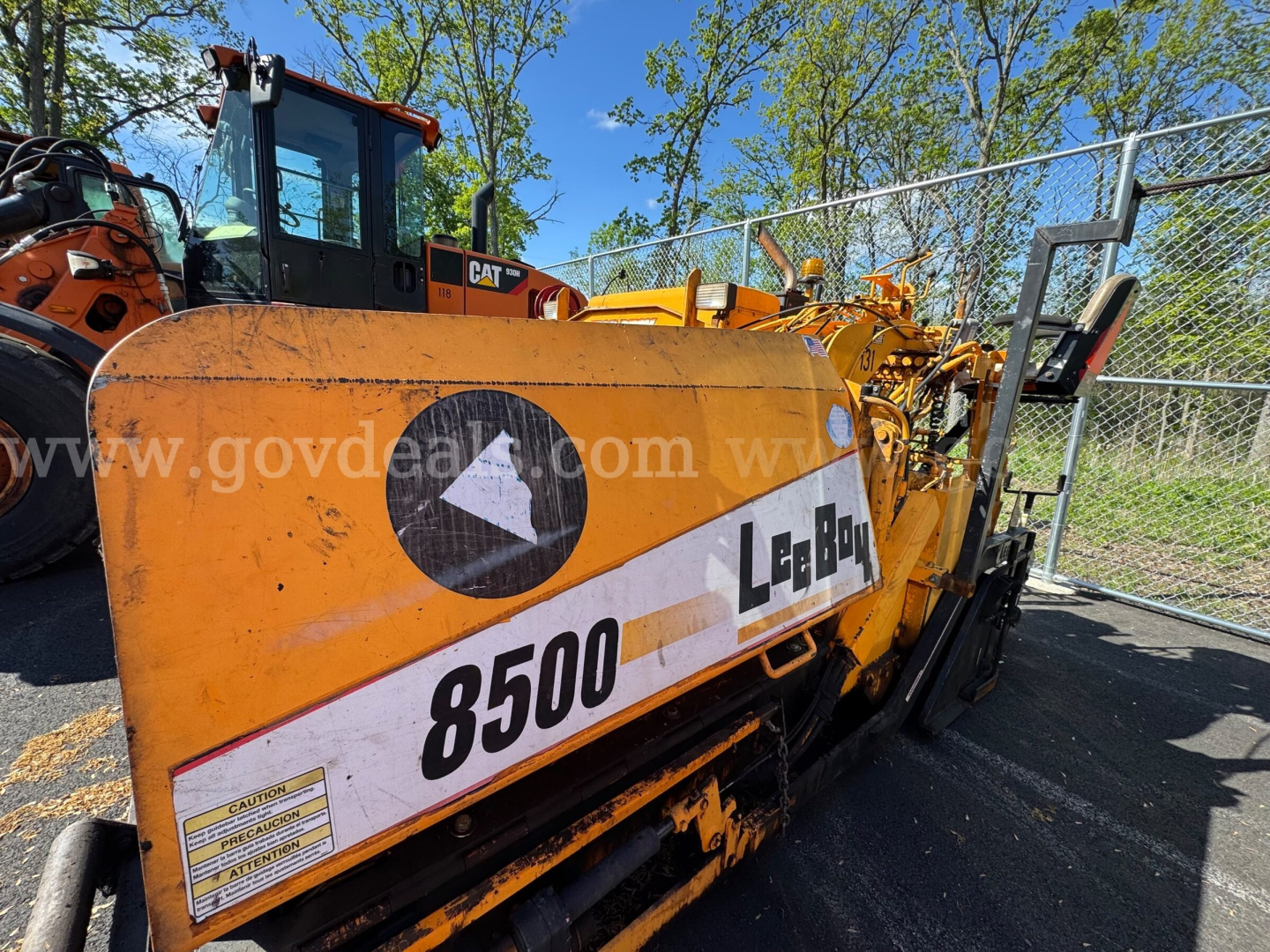 Mack asphalt paver with yellow body showing model 8500 and L-2B9 markings, viewed from side with operator cabin and chain-link fence in background.