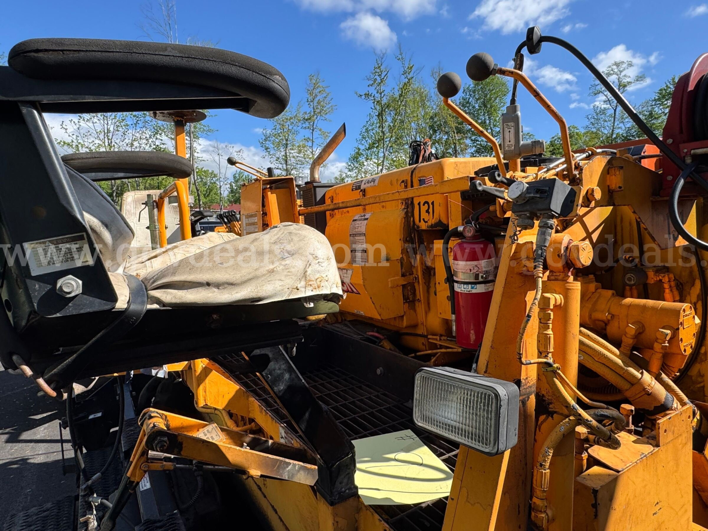Orange Mack asphalt paver with operator seat and control levers, side view showing hydraulic components and warning lights.