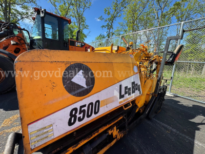 Mack asphalt paver with yellow body showing model 8500 and L-2B9 markings, viewed from side with operator cabin and chain-link fence in background.