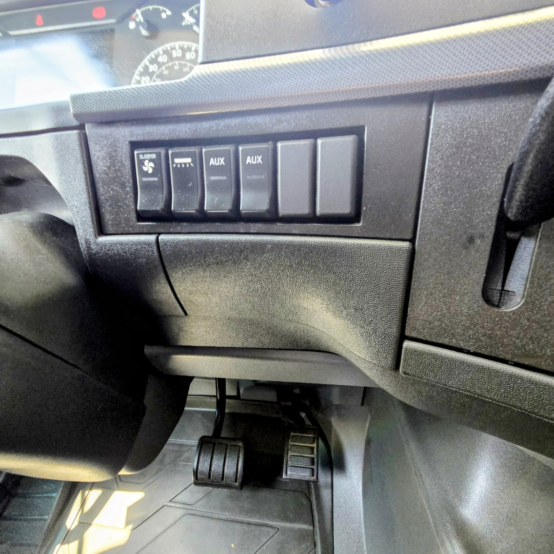 Dashboard area of a Volvo VNL truck showing switch panel with AUX and other controls, foot pedals visible below in-driver side cabin.