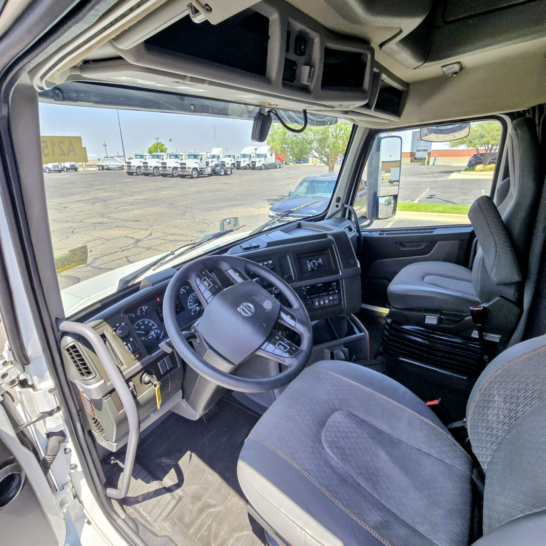 Volvo VNL64T860 truck cab interior showing steering wheel, dashboard, and passenger seat from driver’s side; visible windshield and side window in a dealership lot.