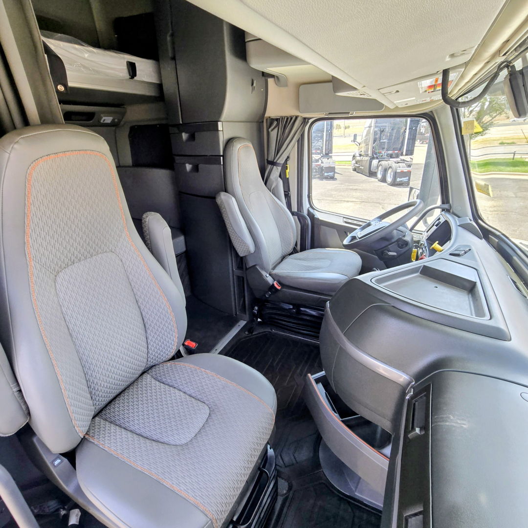 Truck cab interior showing driver's seat and passenger seat with center console, steering wheel, and dashboard in a Volvo VNL64T860.
