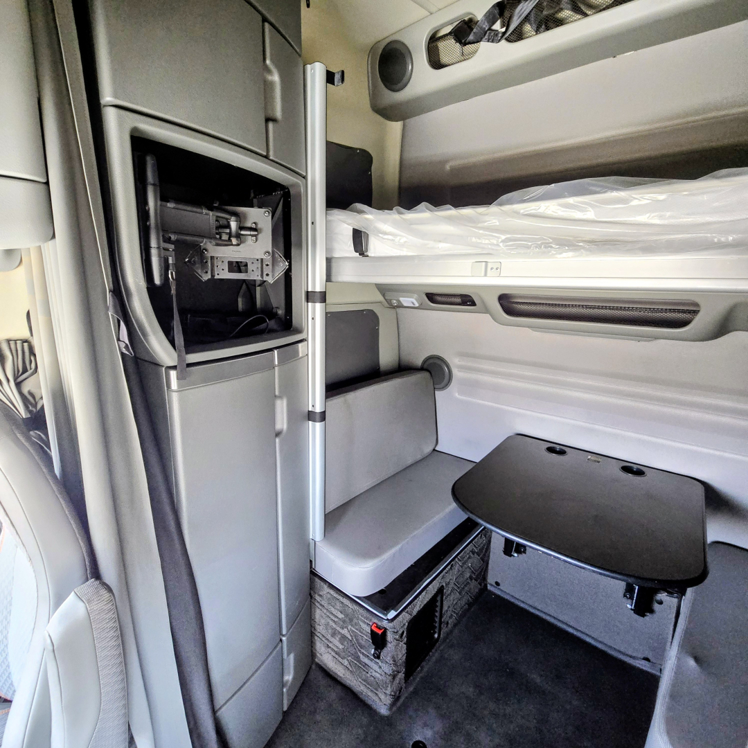 Interior cab of 2022 Volvo VNL64T860 sleeper shows bunk area, storage drawer, and a foldable table near a small cabinet. Visible components include a vertical cab storage panel and seating area.