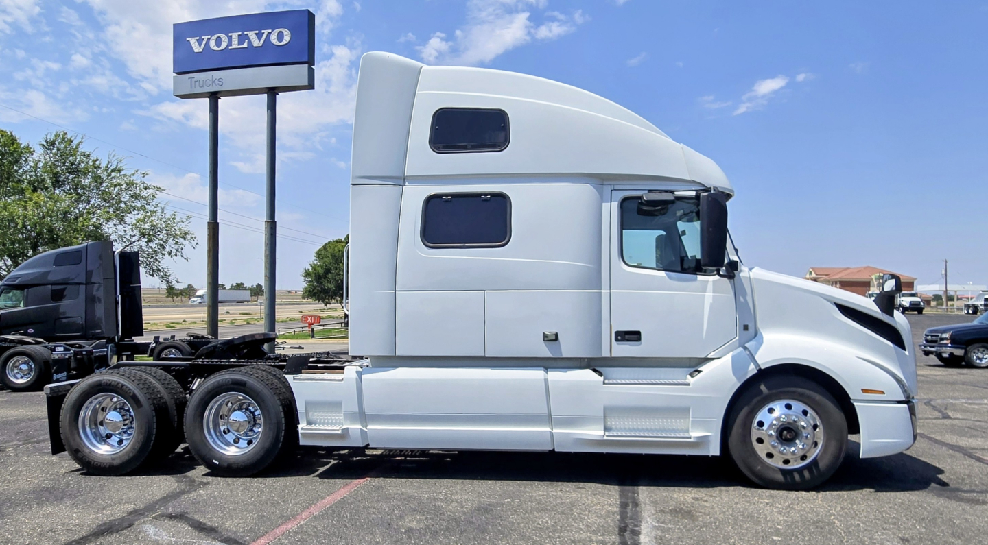 Volvo truck tractor in a lot with a Volvo sign, side view shows sleeper cab and dual rear axles.