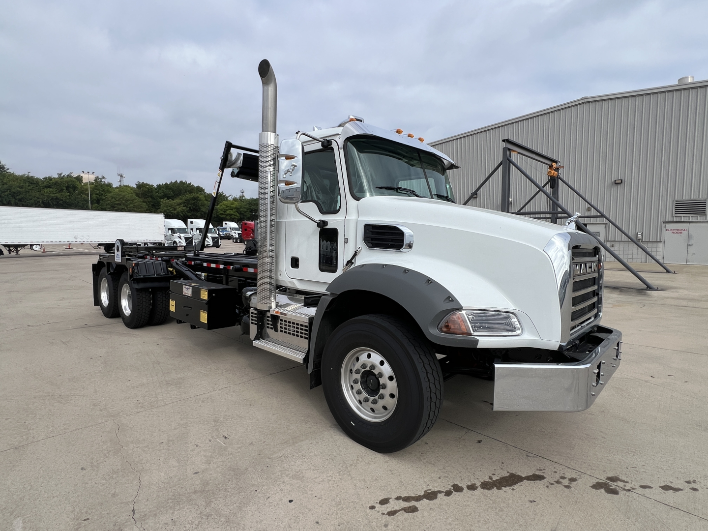Mack GR64B roll-off truck in a dealership lot, white cab, large chrome bumper, tall exhaust stack, flatbed with equipment, front grille with Mack emblem, overcast sky.