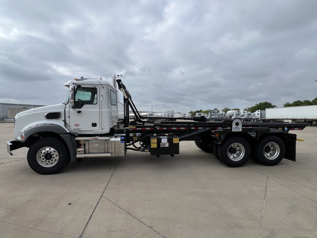 New 2027 Mack GR64B roll-off truck side view showing long flatbed with hydraulic hooklift and dual rear axles, VIN 1M2GR2GC0VM058606, stock 73092.