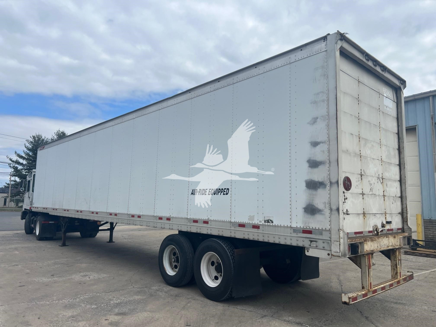 Trailer side view showing Great Dane sheet and post roll door trailer with dual rear axles, light gray sides, and logo of two flying geese. Visible text near logo reading 'LARGE LOW' or similar but not fully clear.