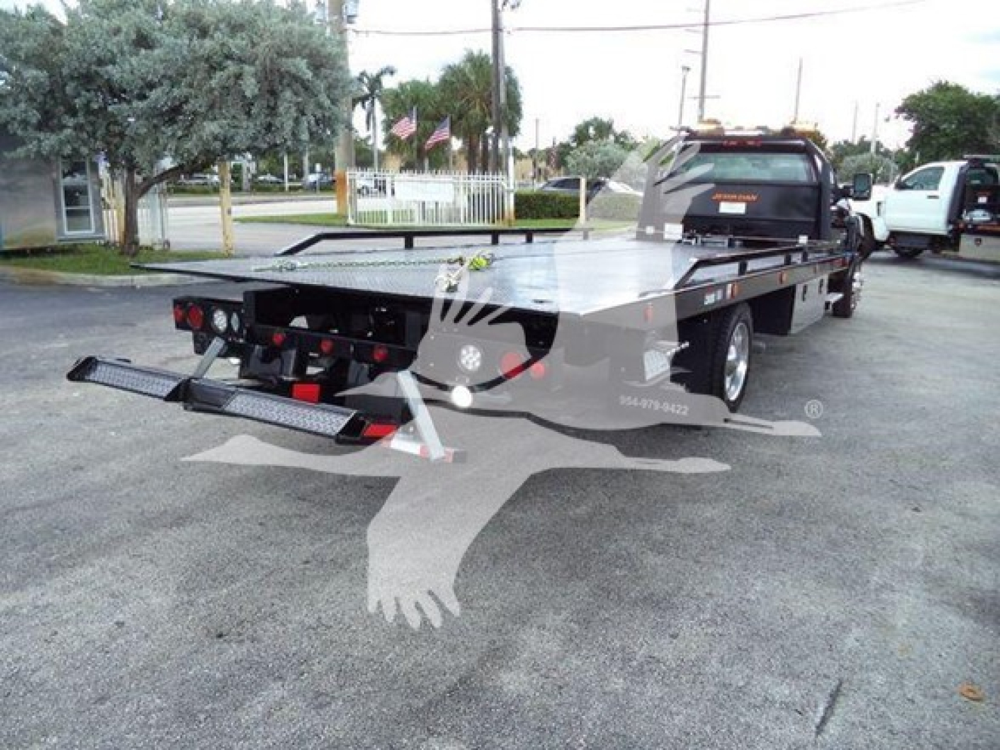 Flatbed tow truck with rollback bed and rear ramps visible, Chevrolet Silverado 6500HD—black flatbed, dual rear wheels, extended cab in background.