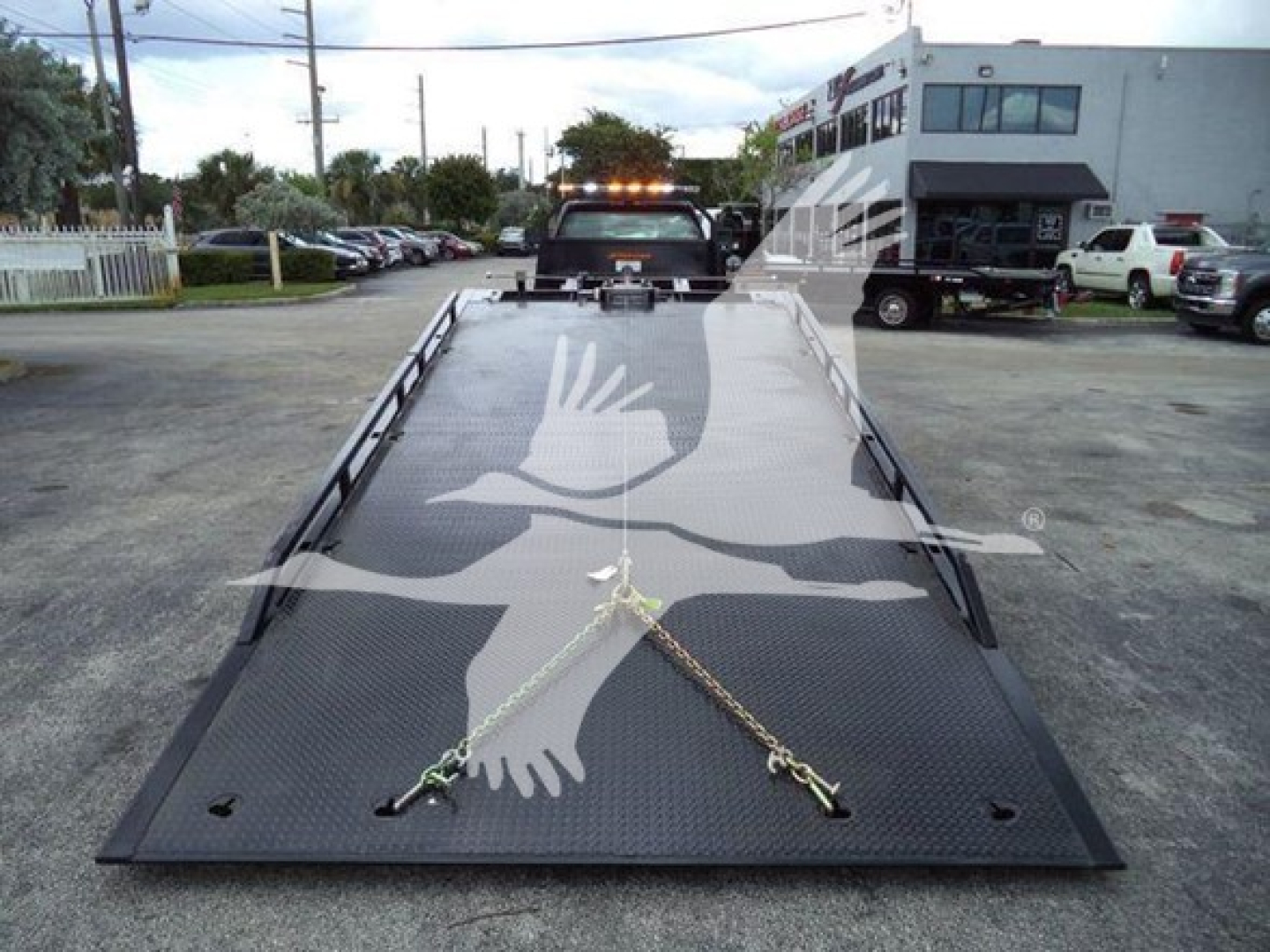 Flatbed tow truck ramp with two chains secured at front, extending toward camera; background shows a service center and parked vehicles.