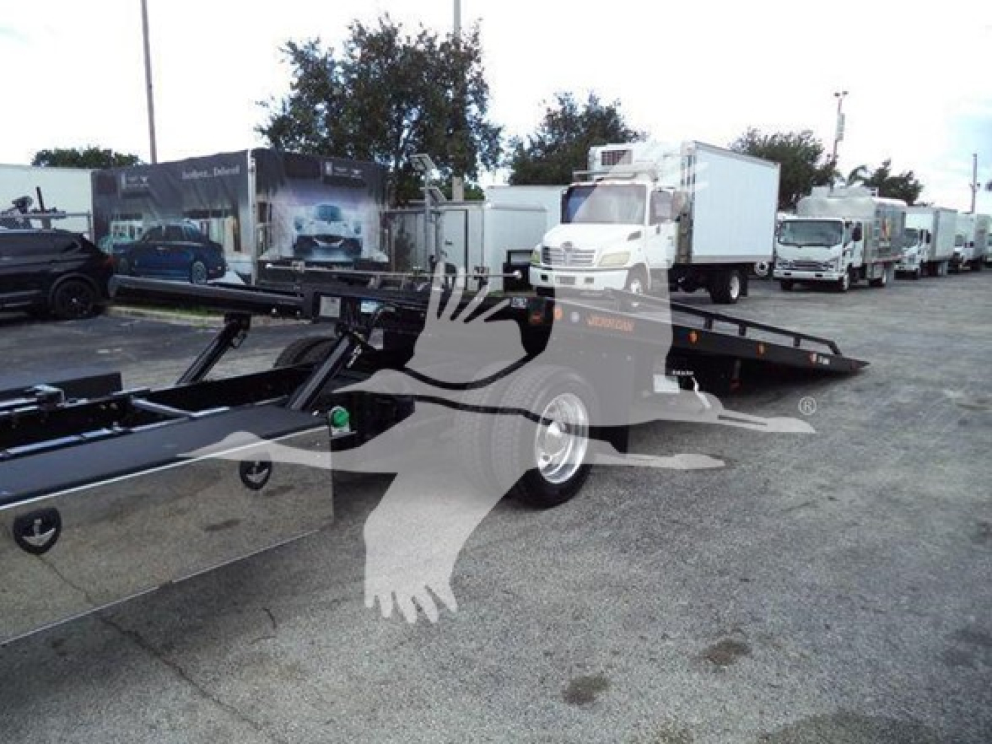 Truck rollback body drop deck showing industrial equipment, black trailer frame with hydraulic components, and line of white trucks in background