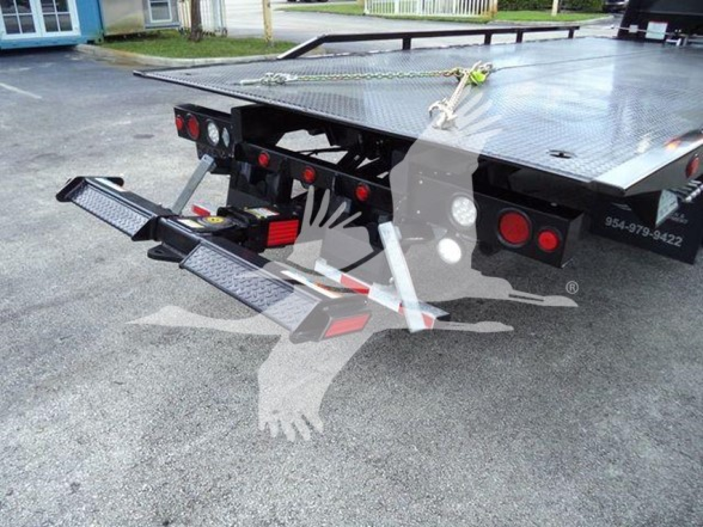 Flatbed tow truck rear with dual drop-down ramps and LED taillights; yellow strap securing front, steel platform, heavy-duty bumper, hydraulic components visible.